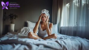 A woman sitting in her bedroom, feeling hot flashes as she experiences menopause. She wipes sweat off her forehead with a towel.